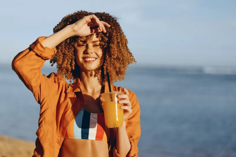 Woman curly hair smiling holding drink beer seaside outdoors wind relaxation, vacation mood, jacket and striped top, hand in hair, holding beer glass, ocean horizon sunlight, warm sea