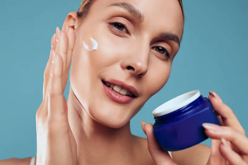 Young woman with clear skin applies white cream from a blue jar, smiling softly against a solid blue studio backdrop, capturing a clean skincare moment with calm, bright vibes.