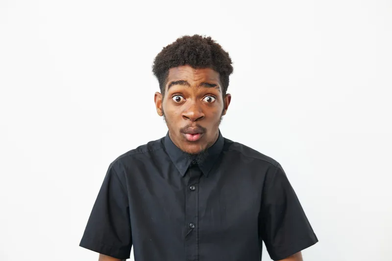 Surprised black man with an expressive face, wearing a black shirt. His wide eyes and mouth open indicate a strong reaction, showcasing curiosity and excitement.