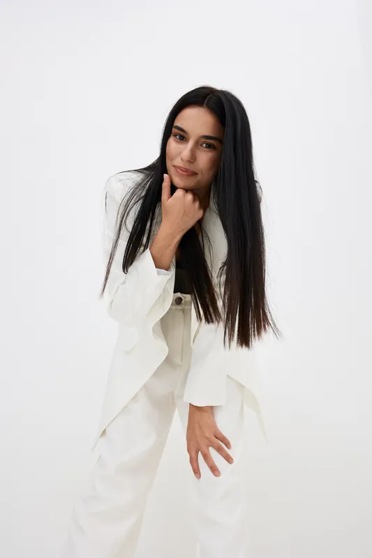 Young woman with long dark hair wearing white pantsuit posing confidently against a plain white background, showcasing modern fashion and natural beauty.