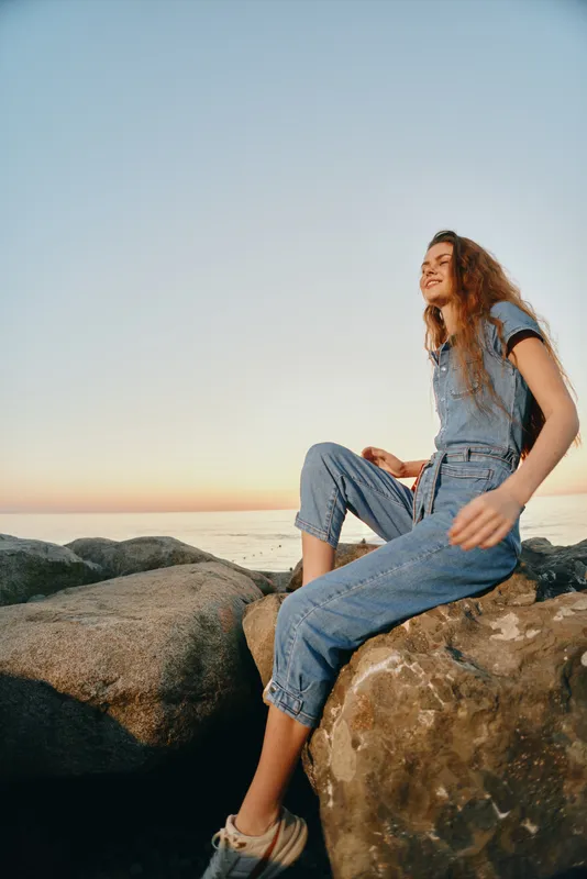lifestyle woman in modern boho-western denim street style sitting on rocks at sunset with relaxed expression, casual outfit, and warm film color tones