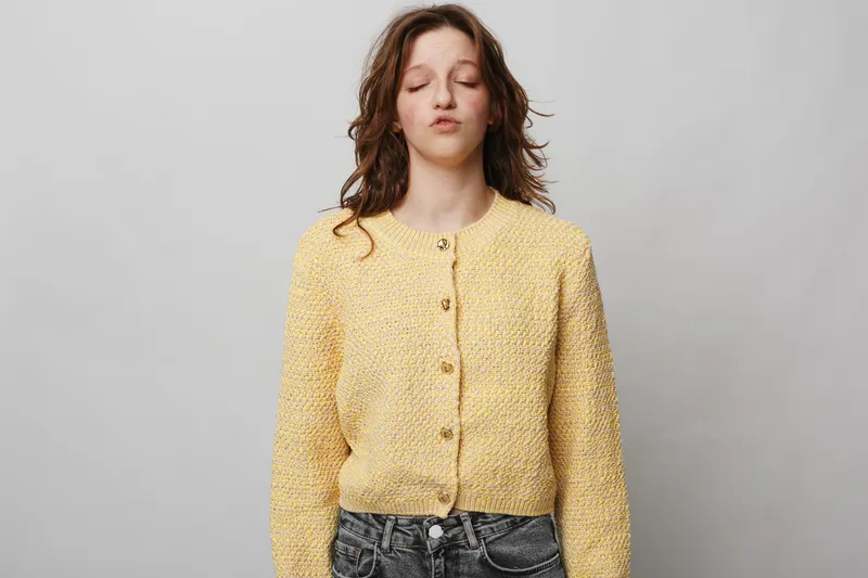 Young woman in yellow knitted cardigan stands with closed eyes and relaxed expression against grey background. Concept of calm, mindfulness, and peaceful moment in casual outfit.