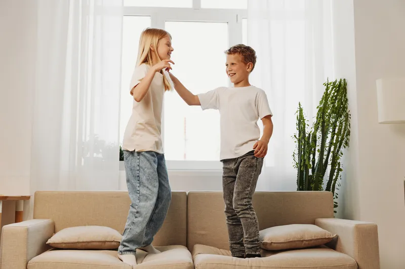 Two happy children jump on sofa in bright living room, smiling and playing together. Concept of joyful childhood and family fun.