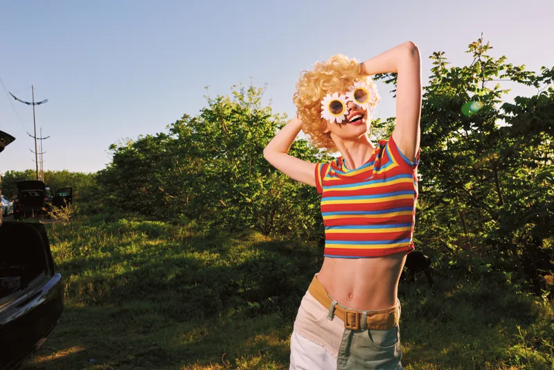 Young woman with a blonde curly wig and flower sunglasses poses outdoors in a striped crop top, radiating retro summer energy and carefree joy.