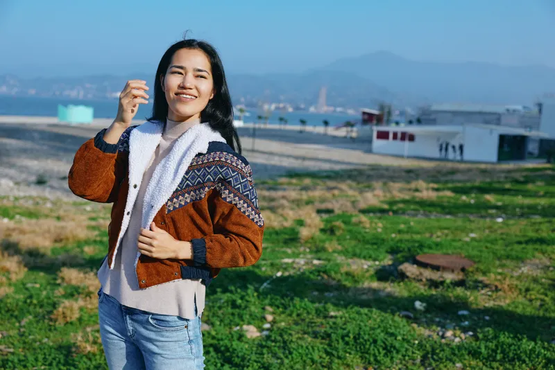 Woman outdoors wearing a knitted jacket and scarf, smiling in a grassy field with distant buildings and hills, capturing casual style, comfort, and a carefree mood on a sunny day