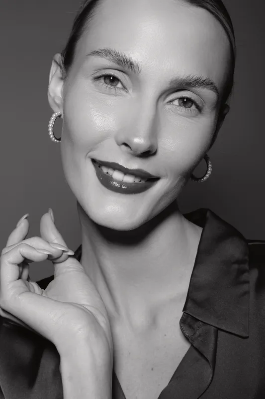 Close-up portrait of a smiling Caucasian woman with fair skin and dark hair styled back, wearing earrings and dark blouse, against a plain background.