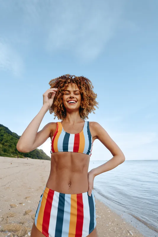 Woman on beach wearing striped swimsuit, smile in summer sunlight, curly hair in hand, relaxed pose near ocean, sandy shoreline and blue sky, vacation portrait for wellbeing and confidence