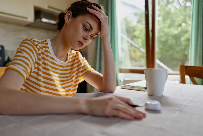 Woman at home sits at a table with laptop and cup, looking stressed and thoughtful, conveying a focused work moment and fatigue during daytime in a calm indoor setting