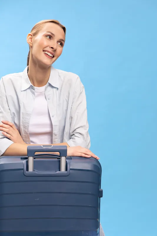Young woman with suitcase smiles looking aside on blue background. Travel concept with luggage, joyful female traveler ready for vacation or business trip indoors studio.