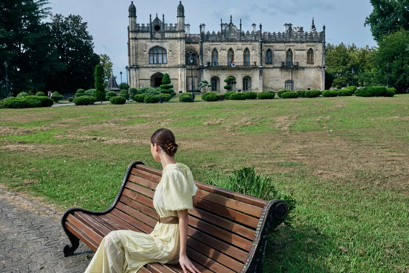 A woman in a pale yellow dress sits on a wooden bench in a manicured park, facing a grand castle like building, evoking quiet elegance and scenic charm.
