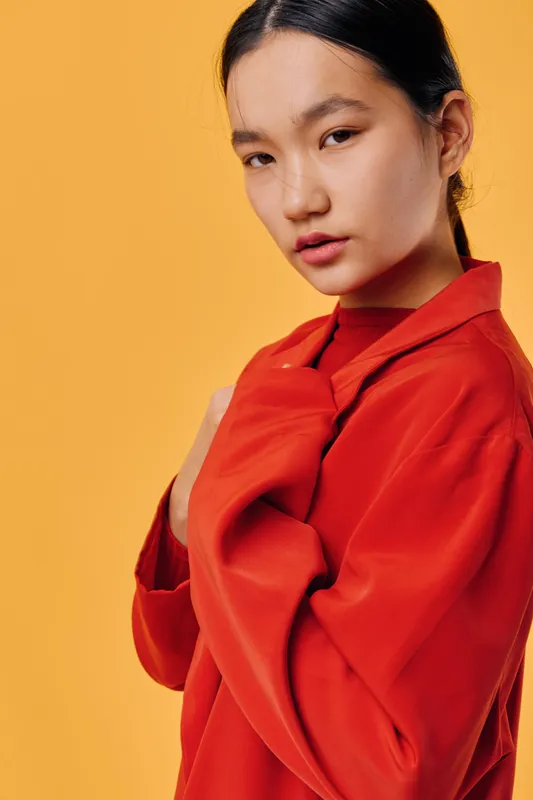 Young woman wearing red coat poses with serious expression on yellow background. Asian female model with natural makeup, looking at camera in studio for fashion portrait.