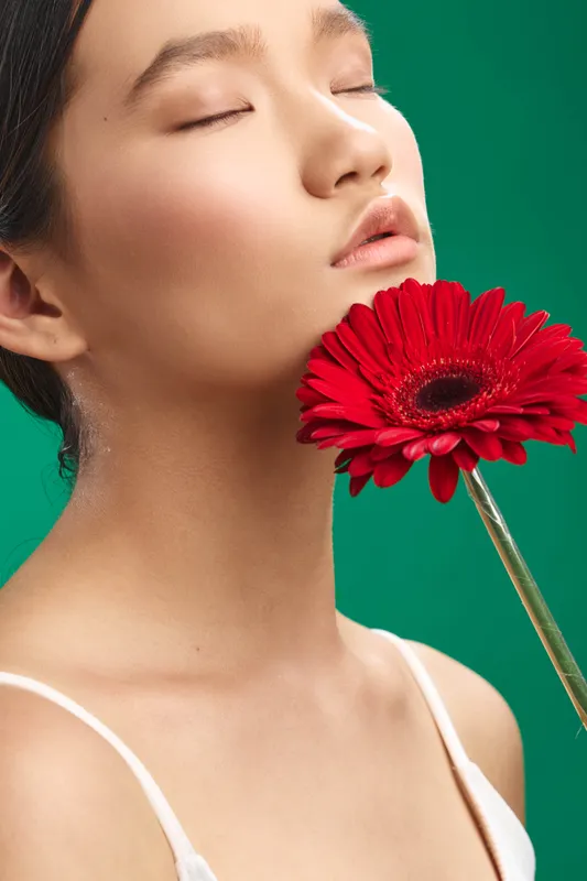 Asian woman in a serene studio portrait with a bright red flower near her lips, soft lighting, and a smooth skin tone creating a calm, intimate mood that feels fresh and artistic.