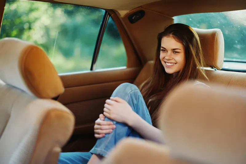 Happy young woman in casual clothing relaxing in the backseat of a car, enjoying a sunny day in nature, conveying a sense of freedom and joy.