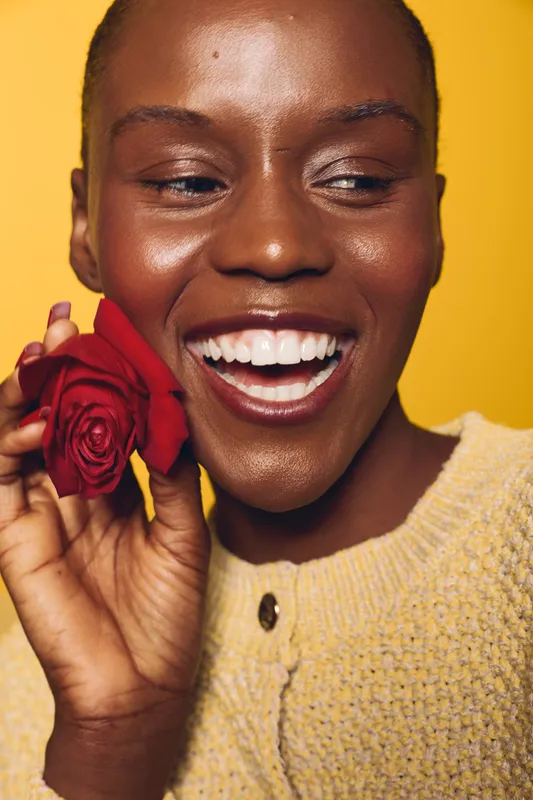 Close-up of smiling dark skinned African woman holding red rose near face, wearing yellow sweater. Bright yellow background highlights joyful mood and natural beauty. Concept of happiness.