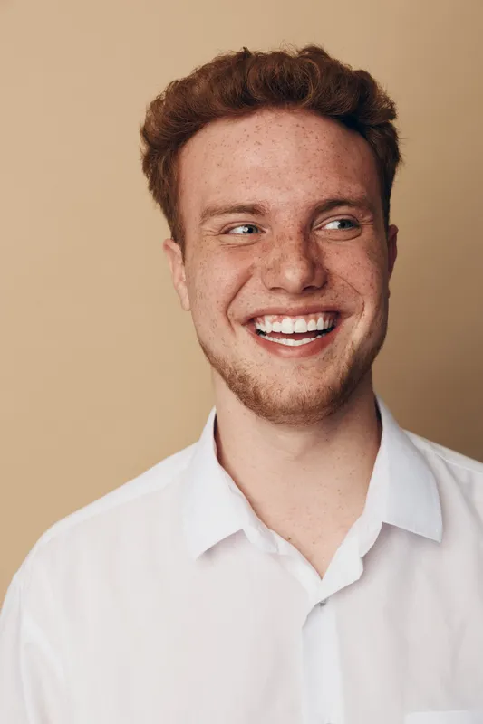 Smiling young man with red hair and freckles wearing white shirt looks to side on beige background. Concept of happiness, youth, casual style, and natural beauty in portrait photography.