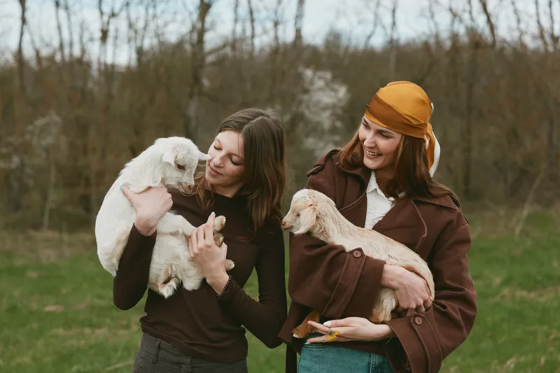 Cheerful women outdoors, holding playful goat kids, warm smiles, rustic countryside backdrop, gentle breeze, natural colors, casual jackets, bonding moment in nature