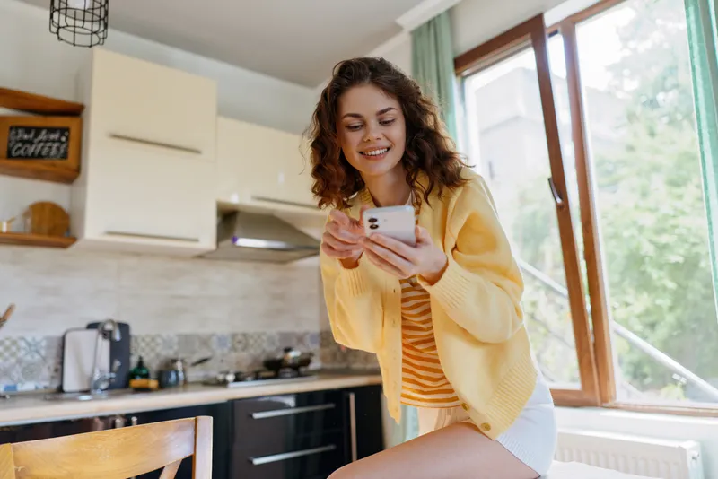 Cheerful woman in a sunny kitchen, looking at her phone with a smile, relaxed pose, casual outfit, natural light, cozy home mood, everyday moment, bright colors