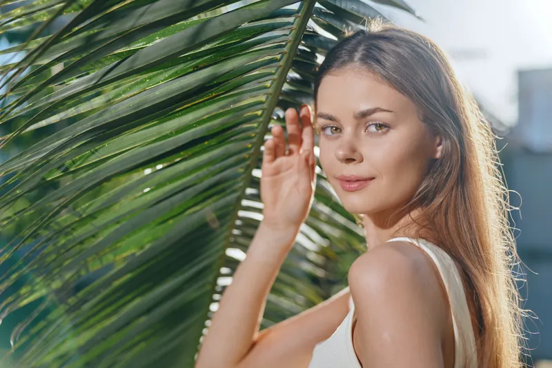 young woman with long hair standing behind palm leaves in sunlight, natural beauty with soft expression, warm summer atmosphere