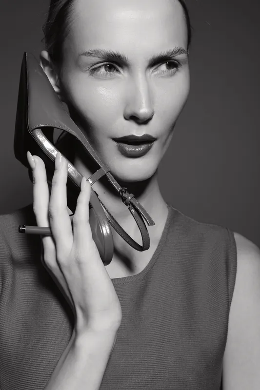 Caucasian woman with light skin holds high heel shoe near face in studio. Fashion concept with elegant female model in black and white.