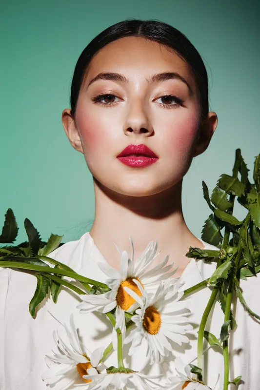 Young woman with glossy lips and subtle blush poses with daisies and greenery on her white top against a soft green background, showcasing creative fashion beauty style.