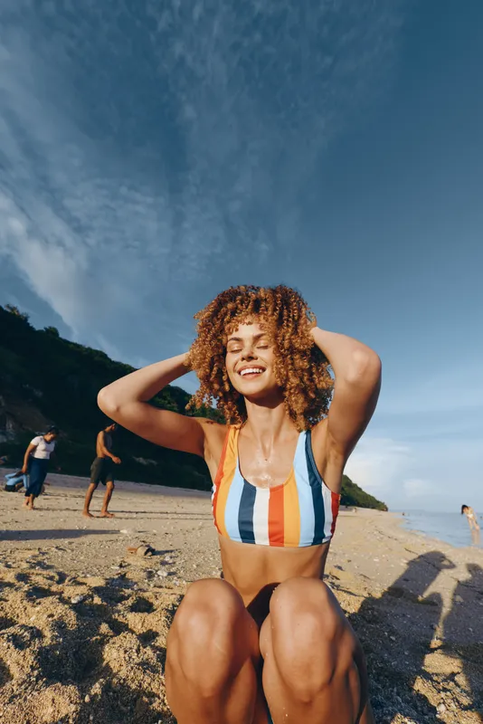 Beach scene with a smiling woman in a striped bikini on the sand, bright sun, blue sky, and relaxed pose, capturing a carefree summer moment outdoors