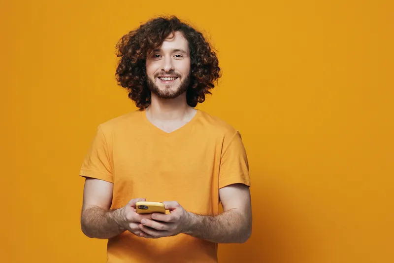 Cheerful young man with curly hair in a yellow t shirt smiling while holding a smartphone against a bright yellow background