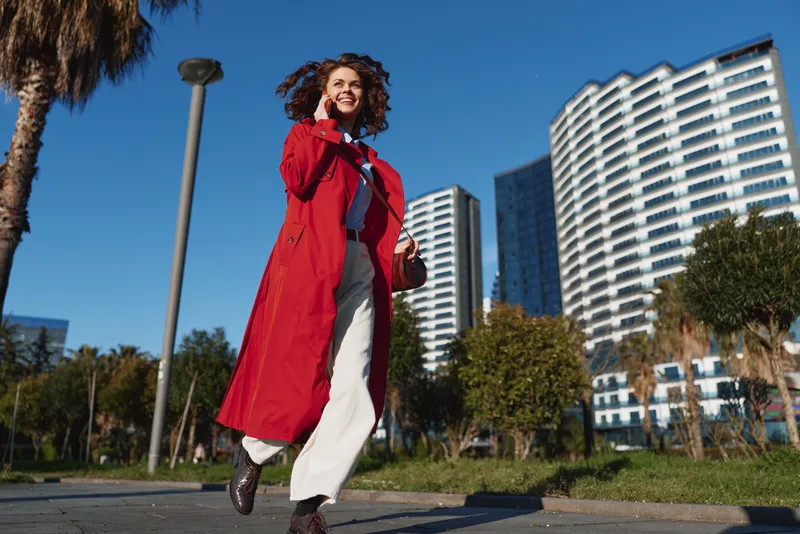 Confident woman walking outdoors in red coat, urban setting with modern buildings, sunny day, smiling face, casual style, positive mood, clear blue sky