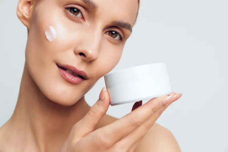 Young woman with clear skin holds a white cream jar near her face while applying moisturizer on her cheek in a bright studio setting with a calm expression.