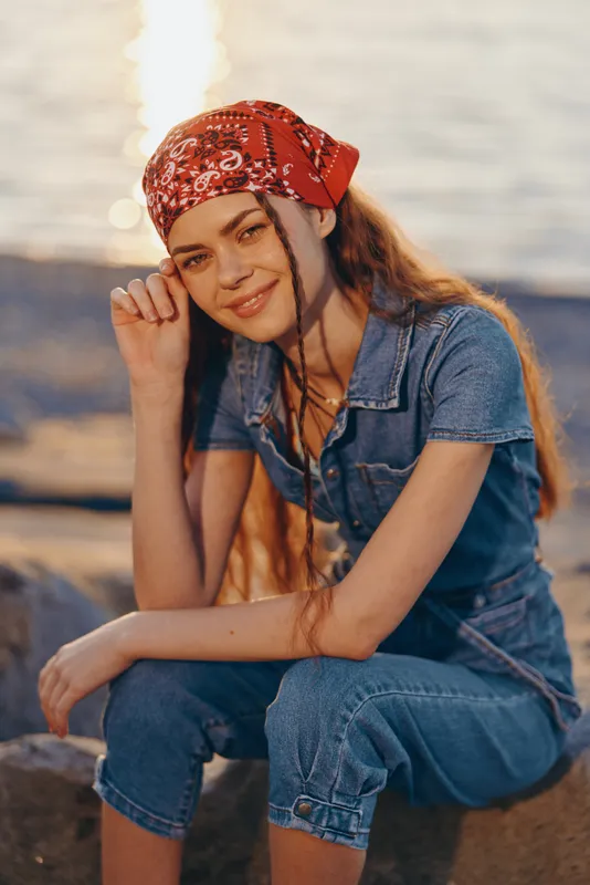 Lifestyle woman wearing modern boho-western denim street style with a red bandana, smiling outdoors near water in warm film color sunlight, casual and relaxed mood.