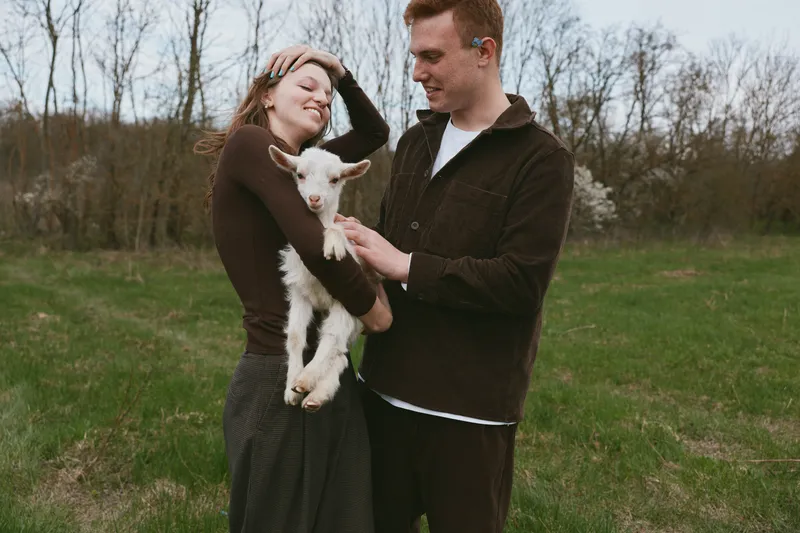 A young couple enjoys a peaceful moment outdoors as the woman holds a baby goat while the man gently touches the animal in a green field during early spring.