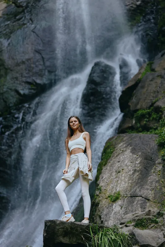 Young woman standing confidently by powerful waterfall wearing white athletic wear surrounded by lush rocks and greenery in natural outdoor setting.