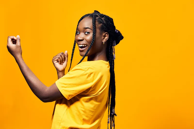 Joyful young girl with braided hair dancing against a bright yellow background, showcasing vibrant energy and happiness in a casual yellow shirt