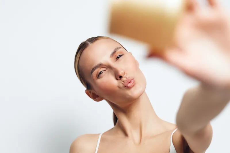 Young woman with pulled-back hair makes a kissing face while taking a selfie against a plain white background in a casual white top.