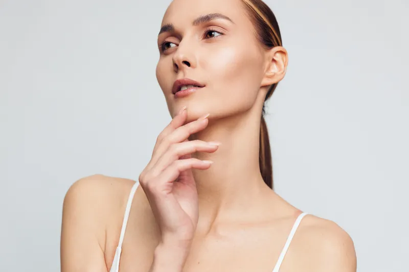 Young woman with clear skin and natural makeup posing with hand near face on light background. Concept of beauty, skincare, health, and natural look. Soft lighting highlights facial features.