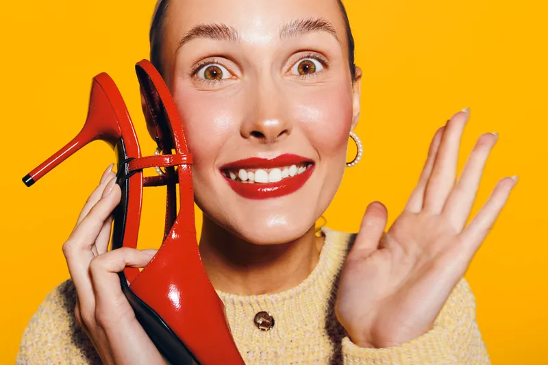 Happy woman with red lipstick holds red high heel shoe near face on bright yellow background. Concept of fashion, style, beauty, and joyful expression in studio lighting.