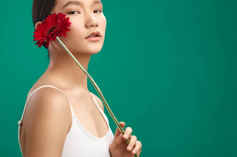 asian woman presents a serene portrait with a bright flower held near her face, set against a solid green background for a clean, artistic and minimal aesthetic.