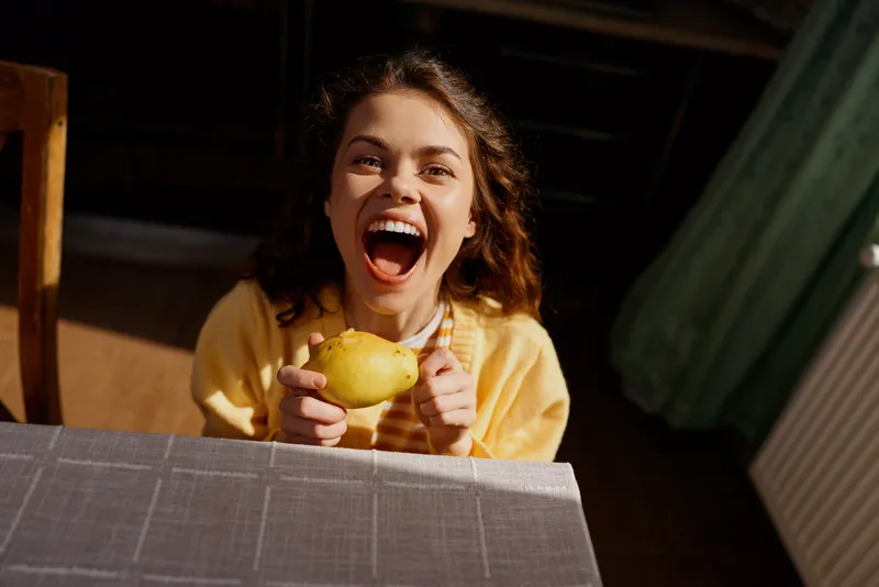 Smiling woman with curly hair holding a pear at a table in a cozy home setting. Casual lifestyle, cheerful mood, and fresh fruit display.
