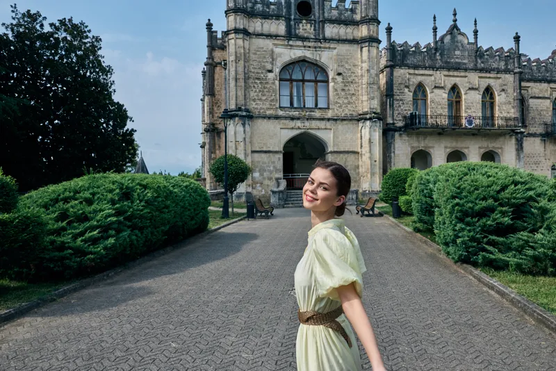 Woman in pale dress stands on a cobblestone path in front of a historic castle, smiling gently as lush greenery frames the grand architecture, and the scene exudes timeless charm.