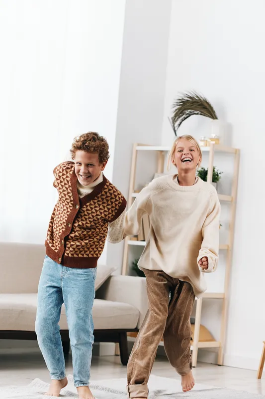 Two children dance joyfully in a bright living room, mid jump with cheerful expressions, showcasing carefree energy, casual outfits, and soft natural lighting.