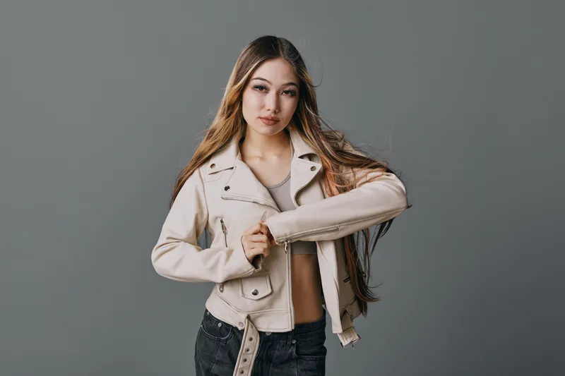 Young woman with long hair wearing a beige jacket and black pants posing against a plain gray background looking confident and stylish.
