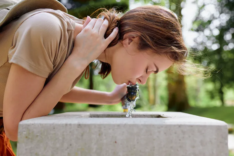 Woman drinking from a public fountain in a park, outdoor summer portrait showing water and hydration as a young casual female leans over a stone basin for refreshing sip.