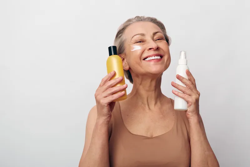 Smiling mature woman holds yellow and white bottles near face with cream stripes, expressing joy and skincare routine concept on light background for beauty and wellness.