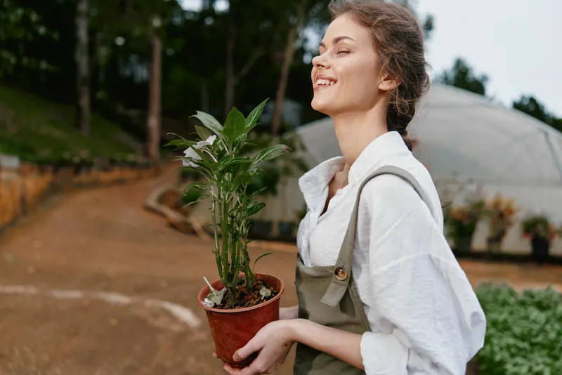 Young woman in a garden holding a potted plant, wearing a light shirt and apron, radiating happiness Perfect for spring gardening themes