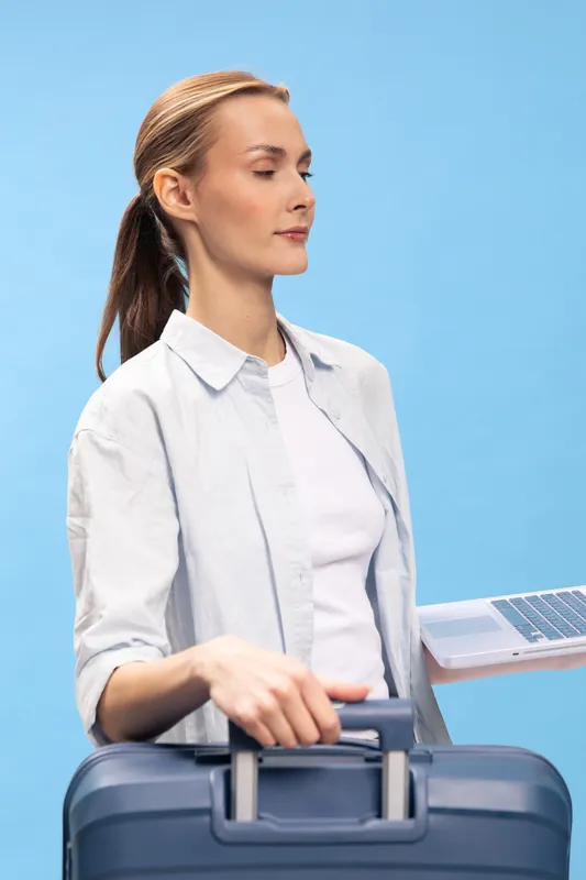 Young woman with ponytail holding a suitcase and a laptop, dressed in casual white shirt and t-shirt, standing on blue background. Concept of travel, business trip, and remote work.