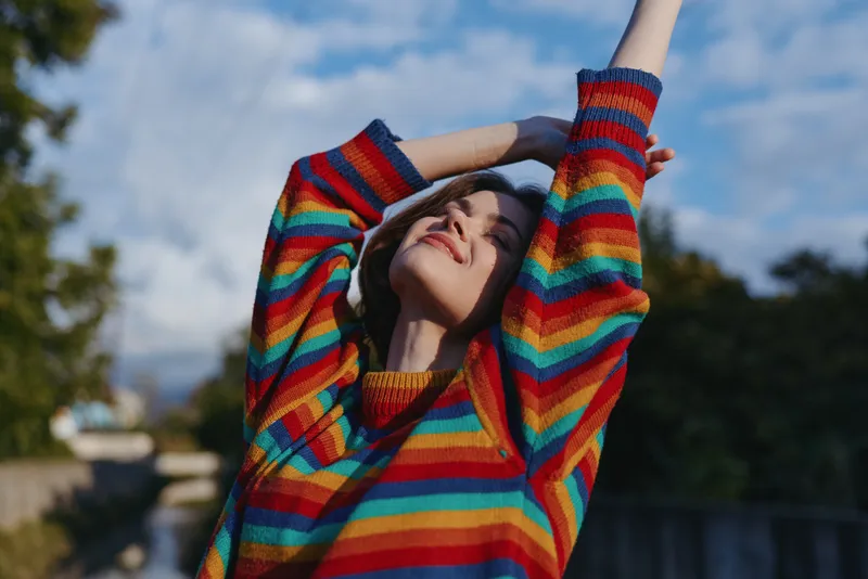 Woman in rainbow sweater stretching outdoors, smiling young adult in her mid 20s with long brown hair, casual outfit and sunlight on face, joyful lifestyle portrait in an urban park.