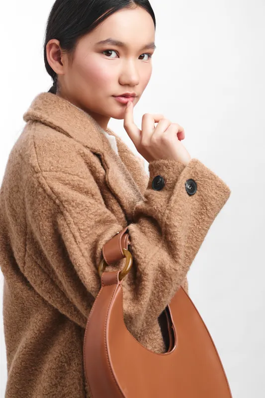 Asian woman wearing a cozy beige coat and carrying a leather bag poses confidently in a studio. She looks thoughtful yet relaxed, conveying fashion sense and calm confidence.