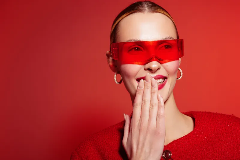 Smiling woman with red sunglasses and red sweater covers mouth with hand on red background. Concept of fashion, style, and modern accessories with vibrant color contrast.