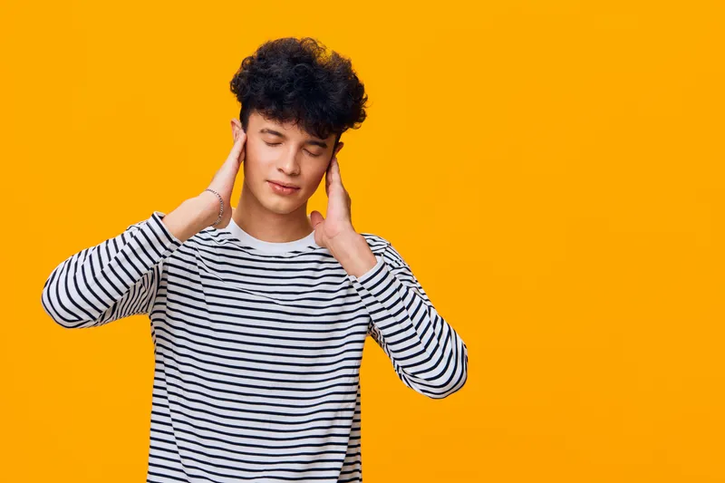 young man in striped shirt with curly hair touching ears with closed eyes against solid orange background, expressing calmness and relaxation indoors