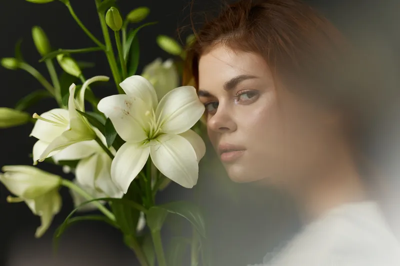 Beautiful young model with brown hair poses with white lilies, showcasing emotions of serenity and elegance against a dark background, perfect for seasonal themes