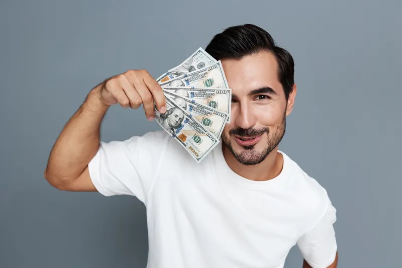 Smiling man holds dollar bills up to his face while posing against a solid gray background, showcasing a joyful expression in a casual white t-shirt.